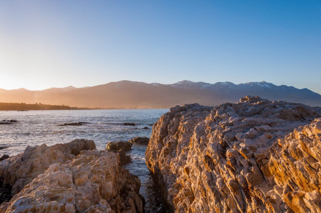 Côte rocheuse dorée par le soleil rasant, face à une chaîne de montagnes enneigées