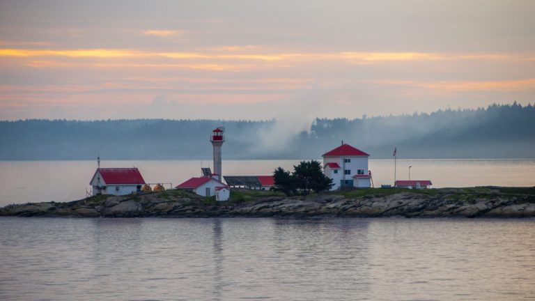 Petit phare et maisons à toit rouge sur un îlot rocheux, brume rose au lever du soleil