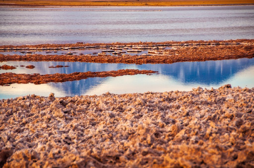 Croûte de sel rugueuse bordant des vasques d'eau reflétant le ciel bleu et rose