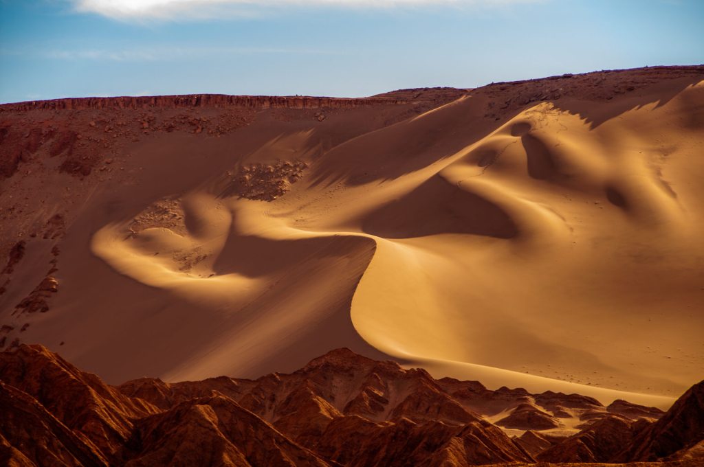 Grande dune de sable clair lovée contre des reliefs de roche rouge découpés