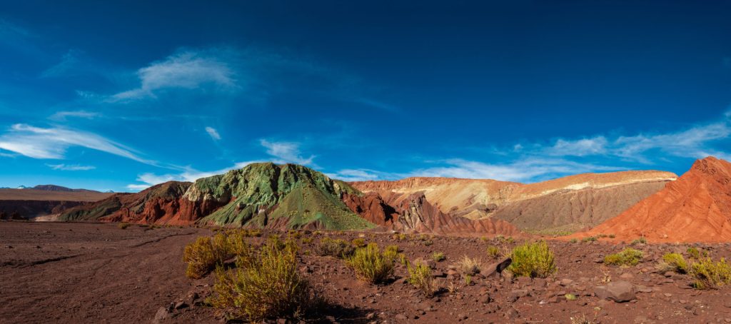 Panoramique de collines minérales aux teintes vertes, rouges et ocres sous un ciel bleu intense