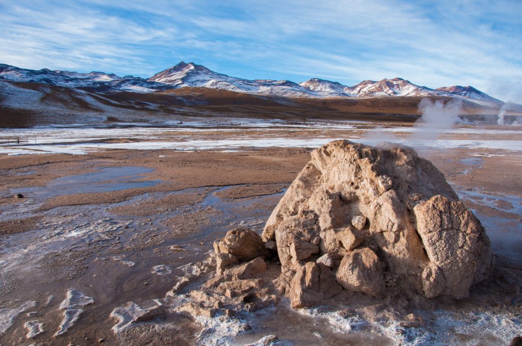 Cône de geyser fumant sur un plateau d'altitude gelé, volcans enneigés à l'horizon