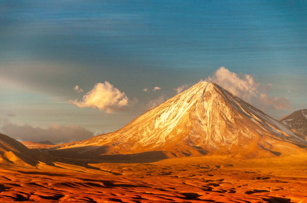 Cône parfait d'un volcan enneigé dominant un désert de sable rouge dans la lumière du soir