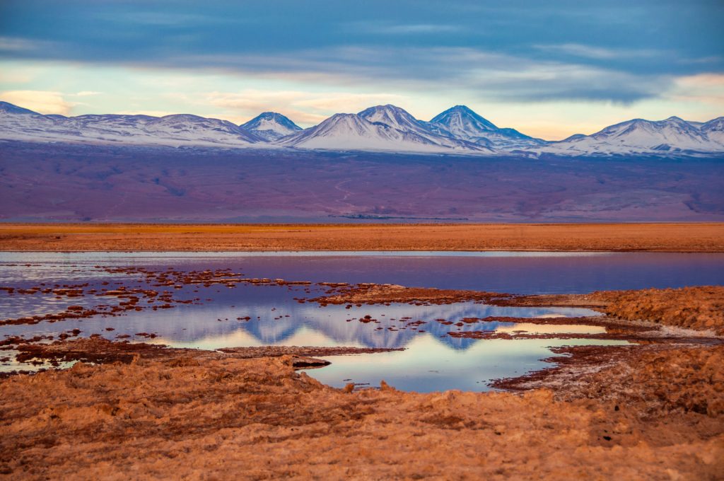Volcans enneigés se reflétant dans les vasques du salar sous un ciel bleu et or