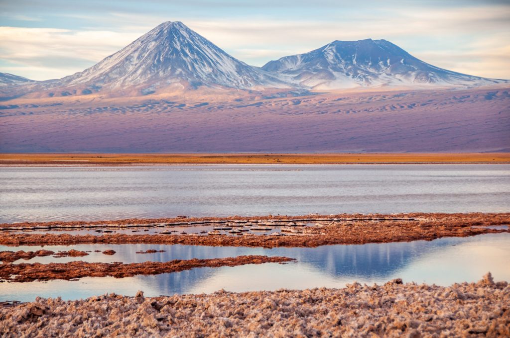 Deux volcans enneigés dominant un salar aux vasques reflétant le ciel pastel