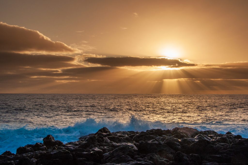 Soleil perçant une bande de nuages et projetant ses rayons sur l'océan, roche volcanique au premier plan