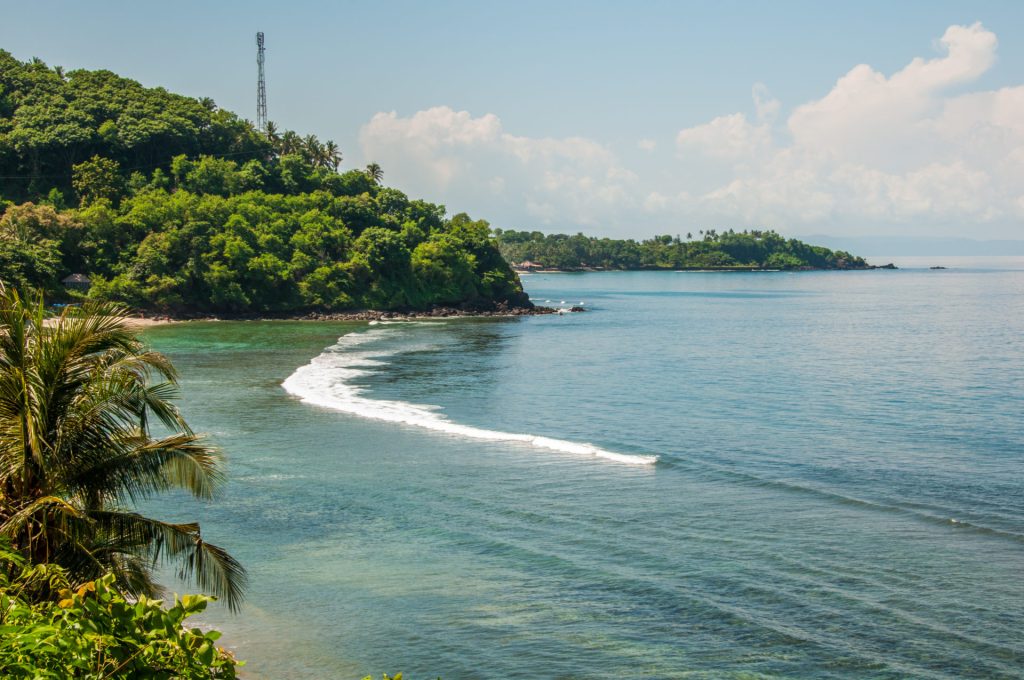 Baie tropicale bordée de collines boisées, longue vague blanche déferlant sur le récif