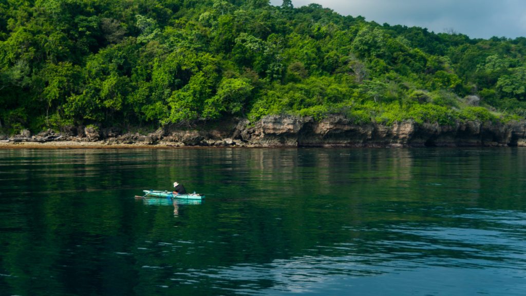 Pêcheur seul sur sa pirogue turquoise devant une falaise couverte de jungle