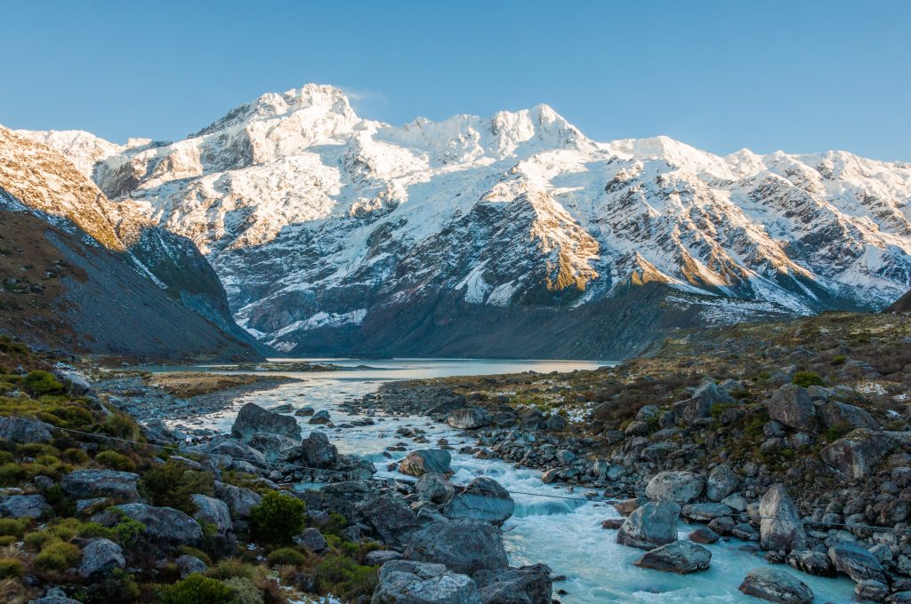 Torrent glaciaire dévalant une vallée rocheuse vers un lac, face au massif enneigé du mont Cook