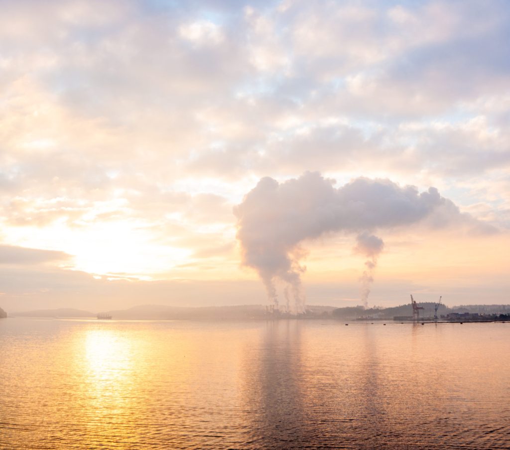 Panoramique d'un port industriel au lever du soleil, fumées verticales et reflets dorés sur l'eau